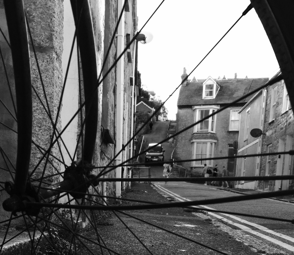 Black and white image: Looking throug bike spokes, up a side street. You can see a car in the distance coming down a steep hill.